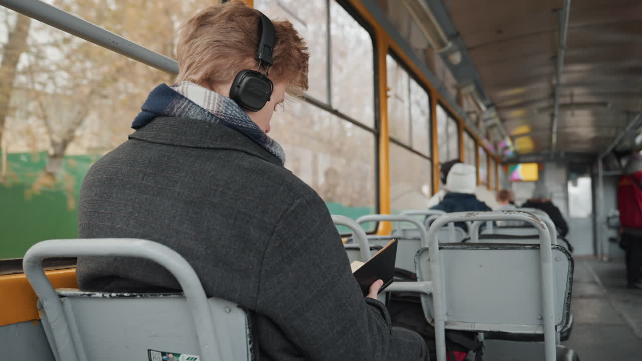 Uomo bianco, creatore, legge sul tram con le cuffie, sfogliando le pagine di un libro. Indossa un cappotto e una sciarpa accoglienti, mentre gli alberi autunnali scorrono fuori. Sedili vuoti, espressione concentrata e pensierosa, un'atmosfera di tranquillo pendolarismo urbano.