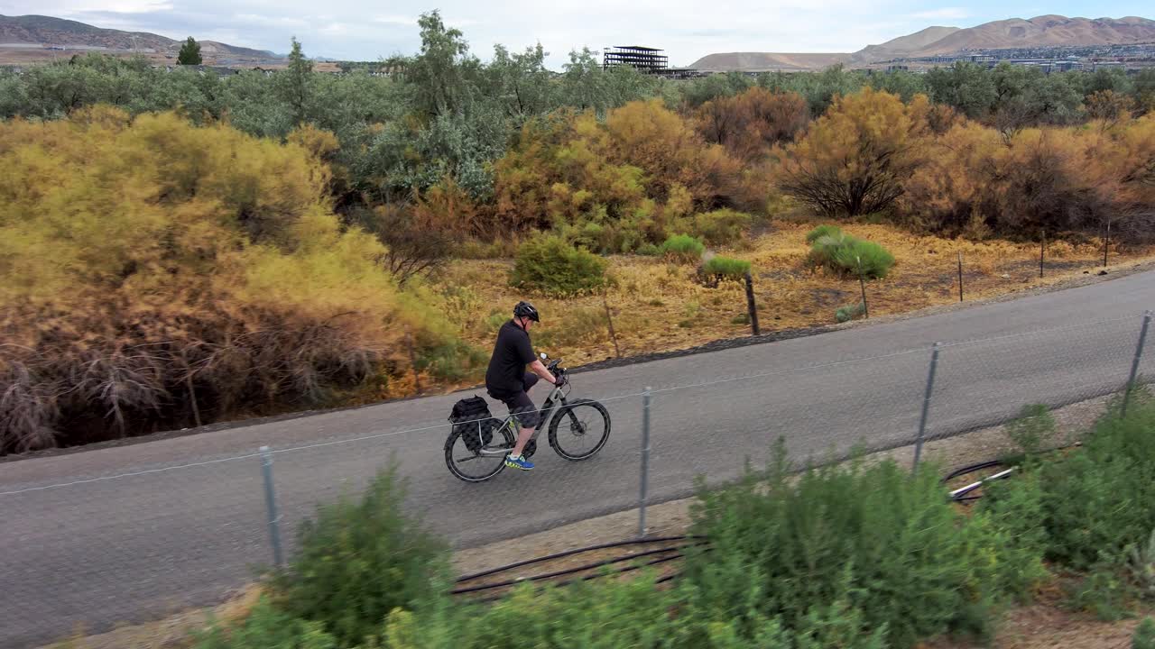 hombre maduro en bicicleta a lo largo de un sendero natural pavimentado con cielo y montañas en el fondo - vista de seguimiento aéreo