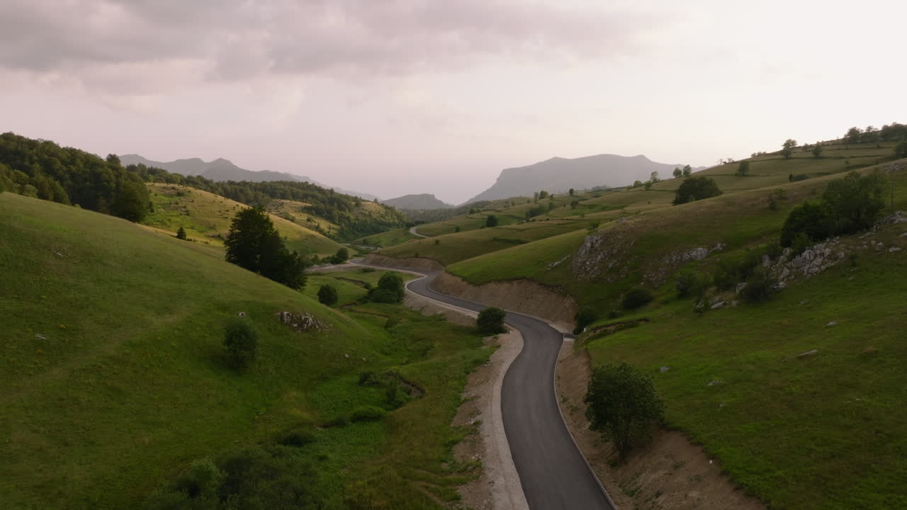 Winding road through a hilly landscape The route cuts through green hills and meadows The sky is overcast Trees dot the scenery