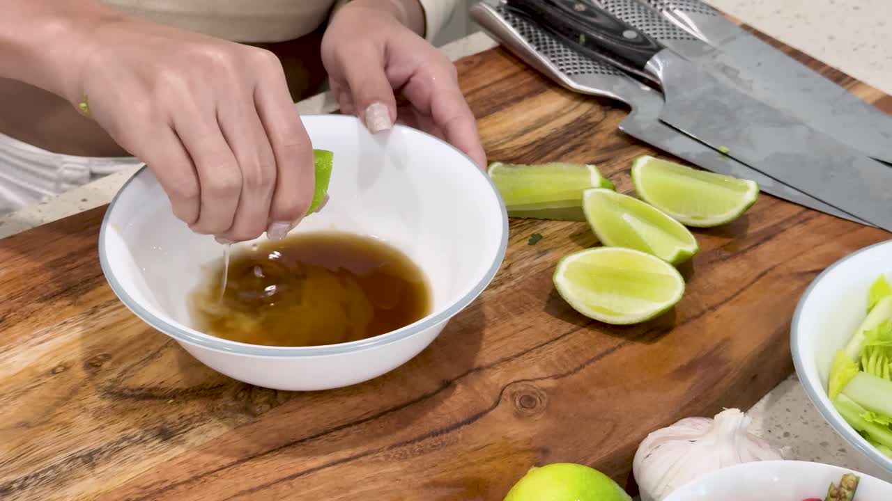 Hands squeeze lime wedges into a white bowl of sauce on a wooden kitchen counter, surrounded by cut limes, garlic, and kitchen knives under bright lighting