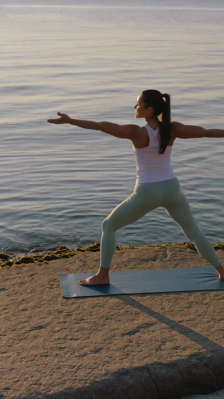 Video verticale: Sole basso che illumina una donna che pratica yoga in piedi su un tappetino blu in riva al mare, canotta, spazio per il testo