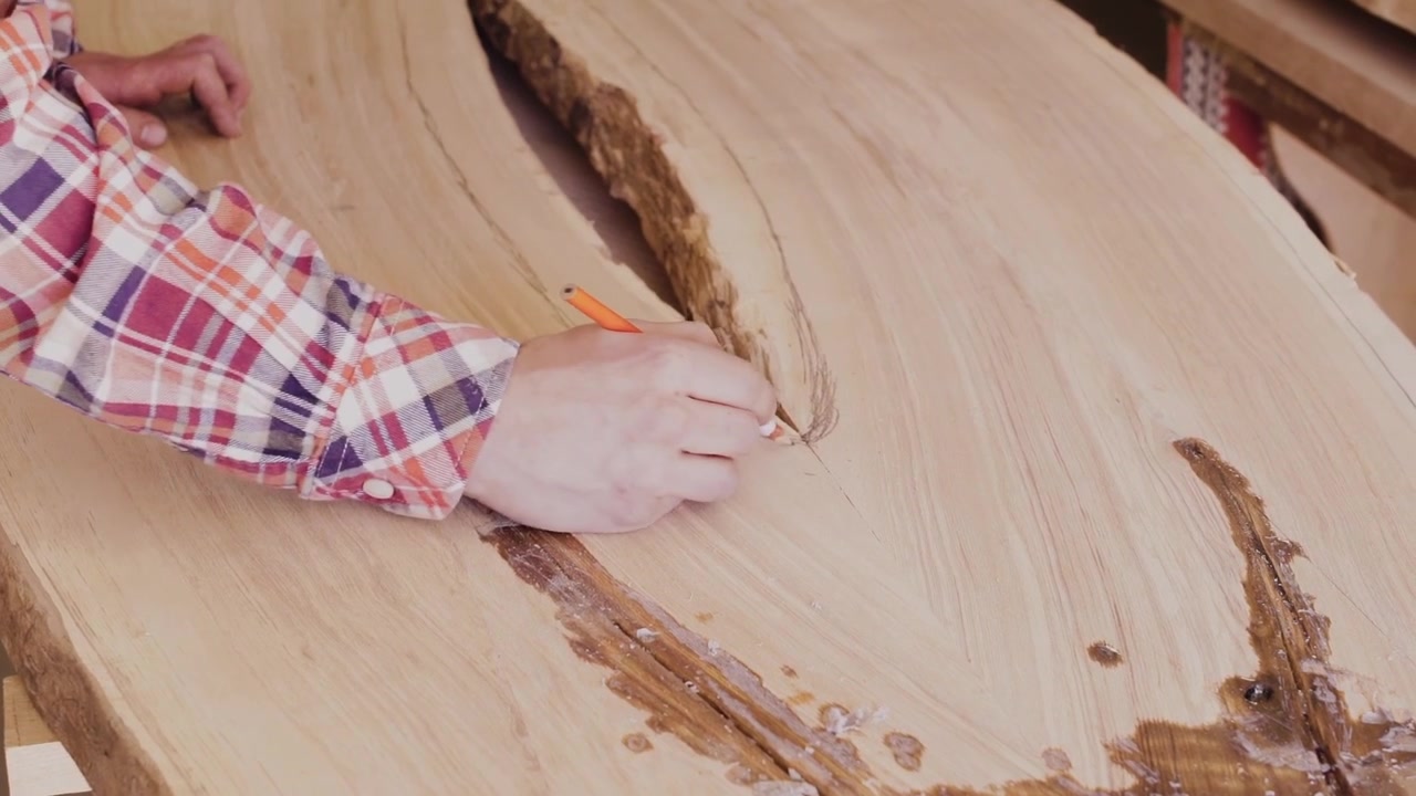 Artigiano che segna la linea di taglio su una tavola di legno con bordo naturale con una matita, preparando il legno naturale per la sagomatura e la lavorazione del legno su un banco da lavoro.