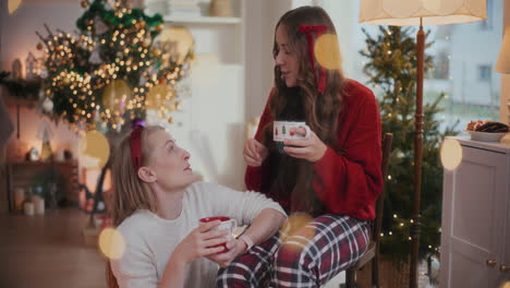 hermanas alegres hablando mientras sostienen tazas de café en la silla durante navidad