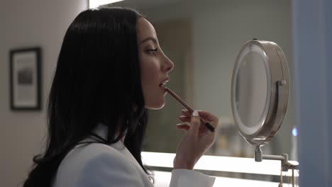 retrato de una mujer hermosa aplicando lápiz labial en la cara, poniendo maquillaje, mirándose en el espejo en casa, preparándose en el baño, vista lateral del perfil