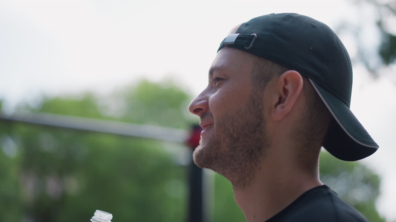 white man drinking water after set, cap back, bottle tilted, droplets on jaw. Athlete replenishes hydration during cooldown at outdoor calisthenics park, smile and relief, sunlight on trees, energy