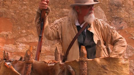 an old man holds a rifle and wears a leather pioneer jacket