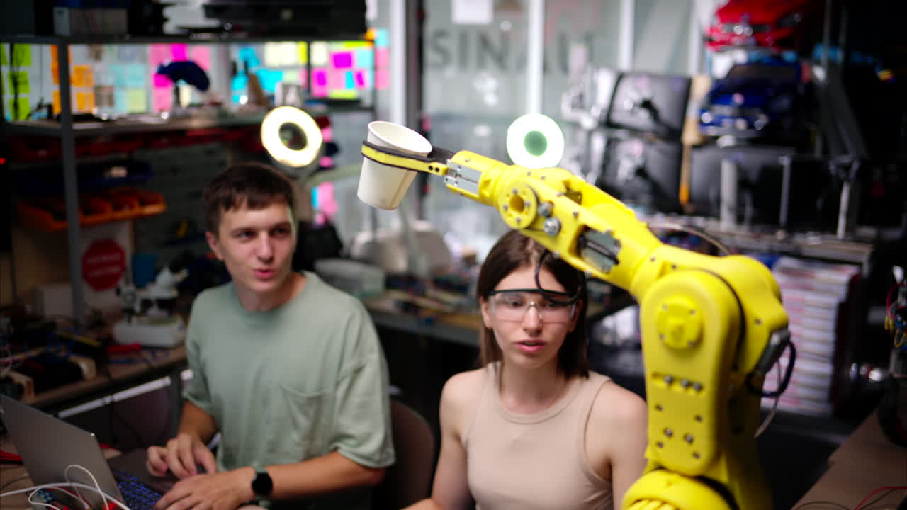 Young happy engineers programming an yellow robotic arm in the workshop to grab cardboard water glass, computer programming training for coffee preparation, celebrating success