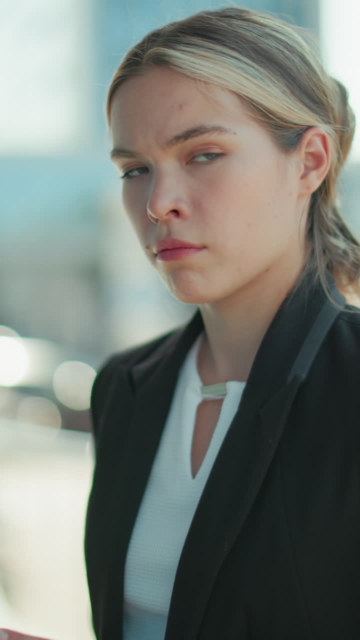 Woman in professional setting operating phone stares at camera with surprised look, dressed in formal attire, standing outdoors with bright urban background of soft bokeh lights and modern buildings