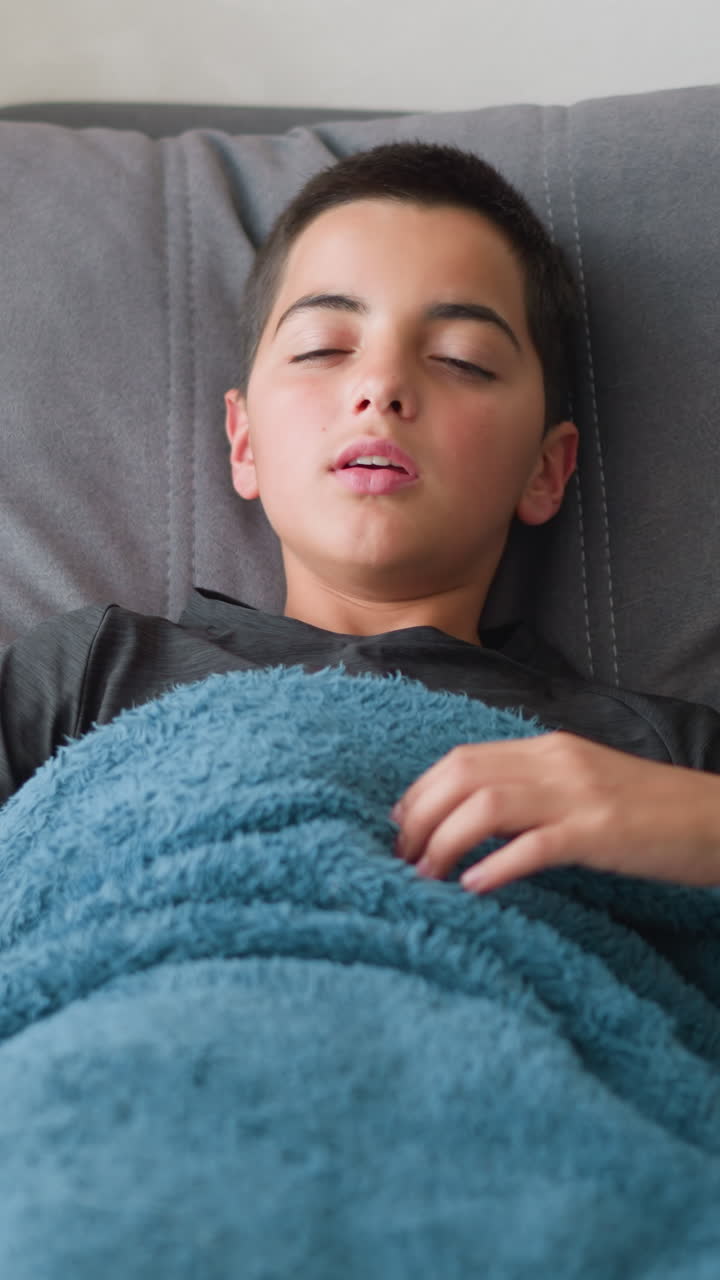 Uncomfortable child sleeping covered in blanket adjusting on couch for comfort with tissue nearby, showing signs of discomfort during rest in peaceful indoor setting
