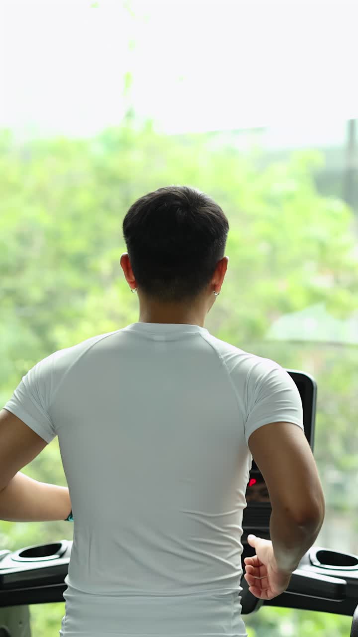 大きな窓と豊かな緑がある明るいジムで、運動能力の高い若い男性がトレッドミルで走っている。