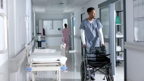 african american male doctor walking and pushing wheelchair in corridor at hospital, slow motion