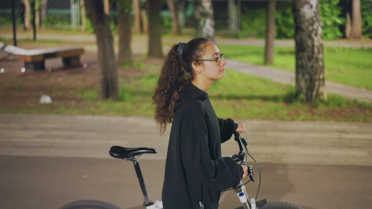 Young Woman With Bicycle In Park, Pushing Bike Along Paved Pathway Under Trees, Calm Midday Atmosphere With Benches And Soft Sunlight, Casual Hoodie And Glasses, Contemplative Expression, Slow Stroll
