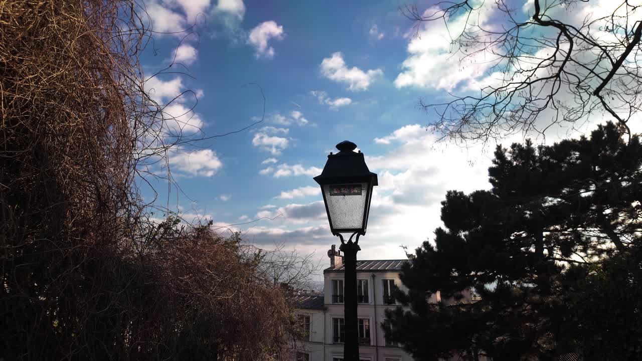 Una farola antigua se erige en Montmartre, París, bajo un cielo nublado. La escena incluye árboles y edificios, capturando el encanto de la arquitectura y el paisaje parisino.