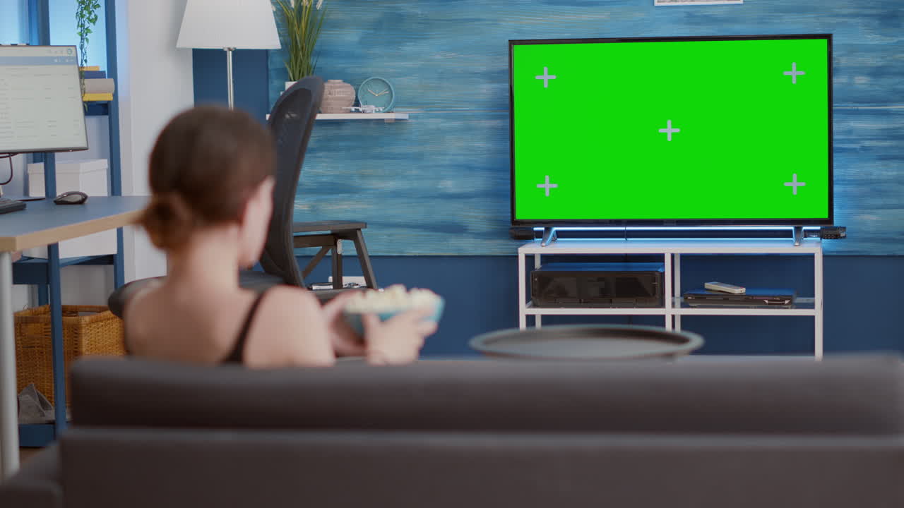 Static tripod shot of young woman watching movie on tv with green screen and eating popcorn