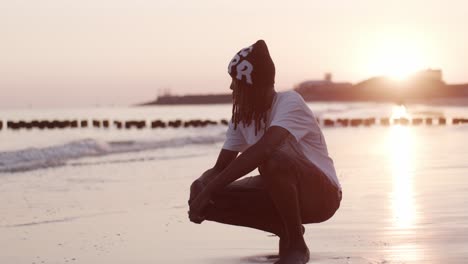 Ein-Afrikanischer-Mann,-Männlicher-Mann,-Modelljunge,-Sitzt-Auf-Kniebeugen-Und-Denkt-Am-Strand,-Meeresufer,-Ozeanwasser-Im-Sonnenuntergang-Nach