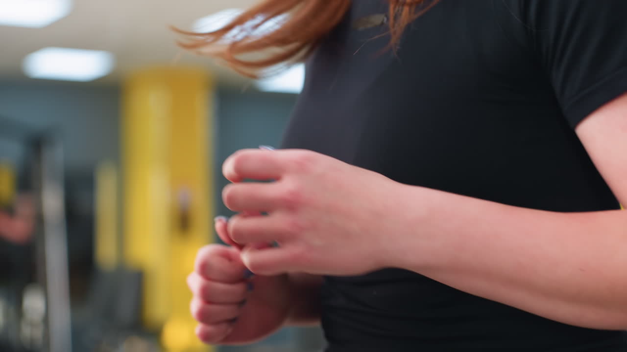 close up of young woman jogging indoors with focused hand movement and visible motion as hair swings during run in bright gym center with colorful background and other people blurred in distance