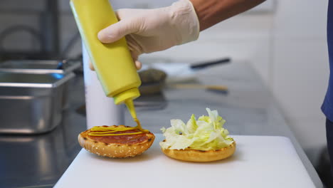 a hand in white gloves pouring sauce on toasted half of the bun. making a delicious hamburger. kitchen in restaurant. front video.