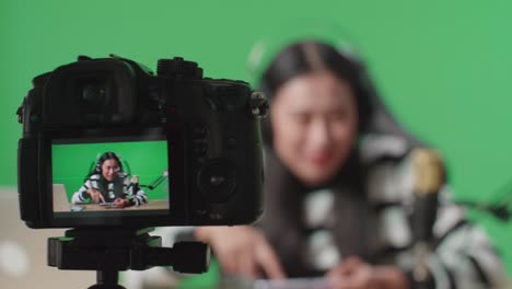 close up de un monitor de cámara grabando a una mujer asiática con auriculares y computadora agitando la mano y luego levantando el dedo índice mientras juega un juego en el teléfono móvil en pantalla verde