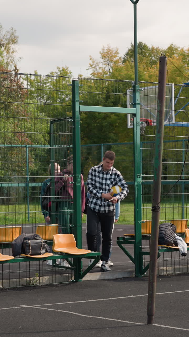 los jugadores de voleibol con bolsas deportivas entran en la cancha al aire libre, preparándose para la carrera de calentamiento con el entrenador, llevan ropa deportiva casual, llevan equipos deportivos, la cancha tiene asientos naranjas y cerca de metal