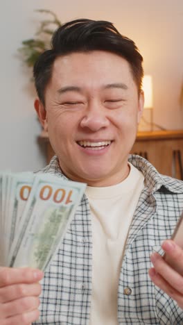 성공한 중년 남자가 집에서 소파에 앉아 스마트폰과 달러 지폐를 들고 있습니다.