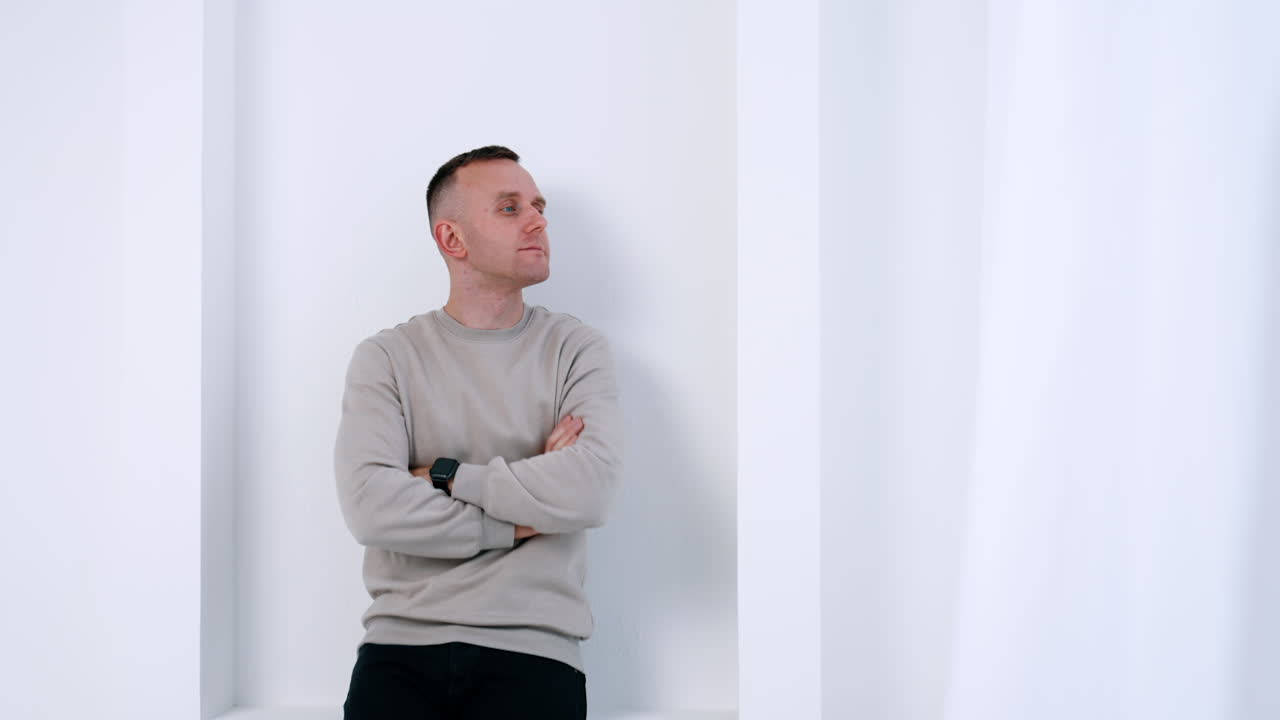 Pensive Caucasian male wearing grey sweater. Man stands at the white wall looking to the side and smiling.
