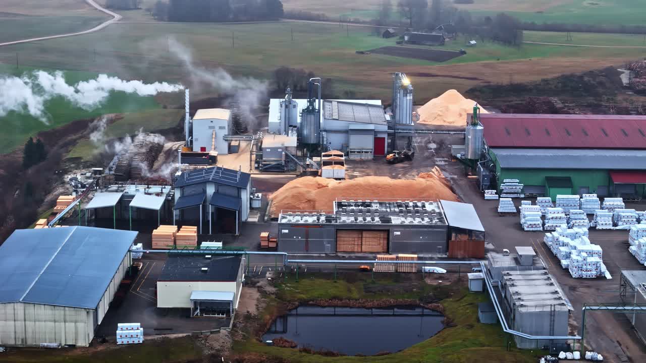 Drone view of a rural industrial processing plant with production buildings, storage areas, steam emissions and material stockpiles. Aerial manufacturing scene in an agricultural landscape
