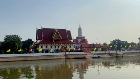 traditional thai temple by river with boats, flags fluttering, clear sky, serene setting, daytime shot, wide angle