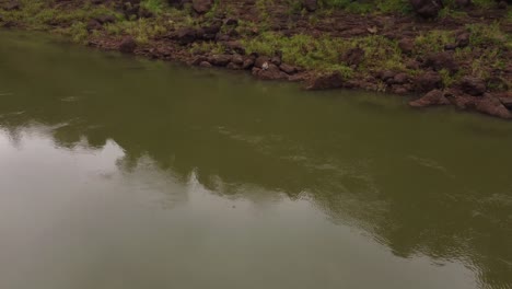 Toma-Aérea-De-Una-Mujer-Sentada-En-Una-Roca-Frente-Al-Verde-Río-Iguazú-En-La-Frontera-Brasileña-Y-Argentina---Aguas-Tranquilas-En-El-Desierto-De-La-Selva-Amazónica