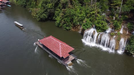 a 4k drone shot of a floating house sailing past a small waterfall in the jungle of sai yok national park in thailand in southeast asia