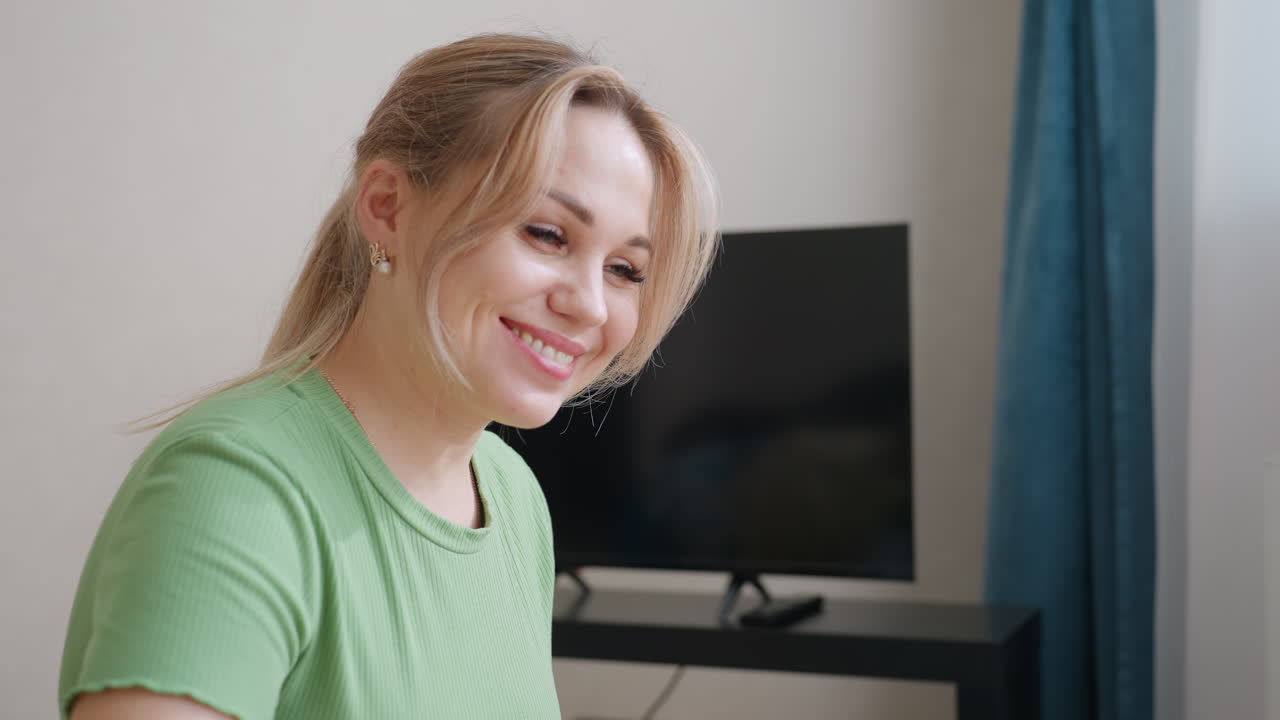 Mother squats close to bed tapping son on nose with gentle touch while smiling warmly to wake him up as television set stands in background creating tender family morning