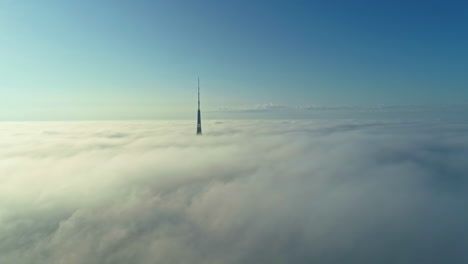 Skyscraper-jutting-through-low-fluffy-clouds,-flying-over-the-clouds-beholding-blue-skyline