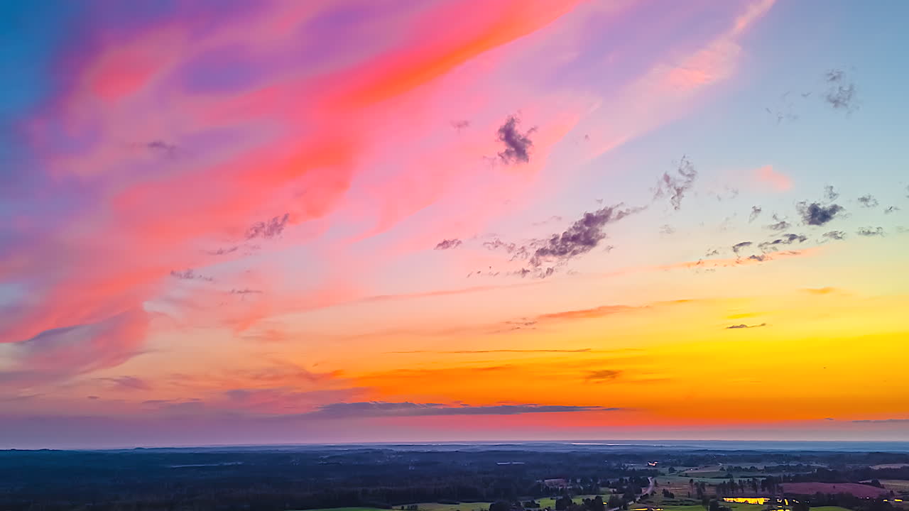 Timelapse de um pôr do sol vibrante com nuvens rosas abrangentes que se dissipam em tons laranja e azul escuro sobre um vasto horizonte rural
