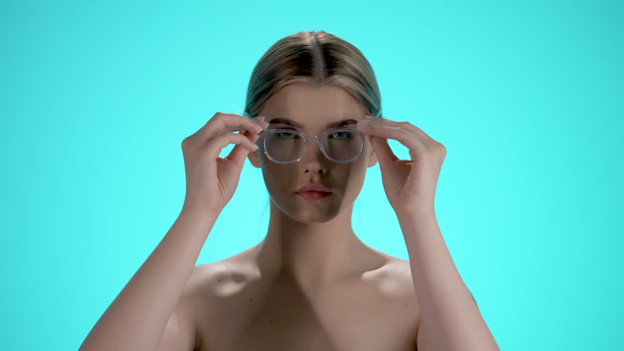 Static medium shot of a pretty young woman pulling her glasses on her head and looking seductively into the camera in front of turquoise background in slow motion