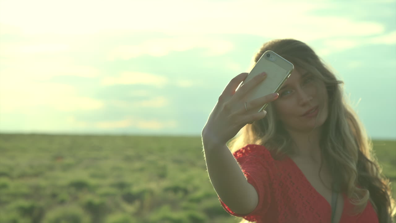 Woman in a red dress taking a selfie with the sun shining behind her hair in a field