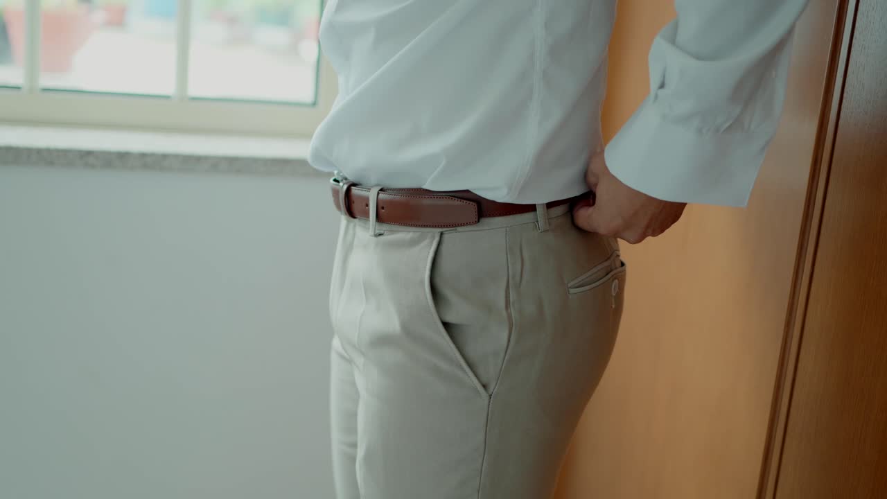 Groom in a white shirt adjusts his brown belt, standing by a window in natural light