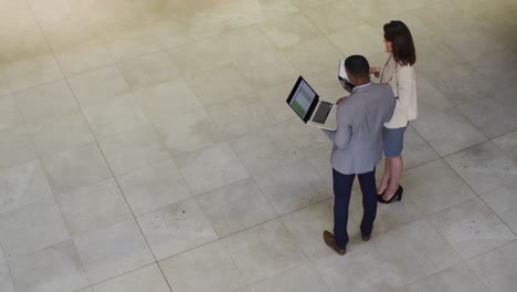 Diverse-businessman-and-businesswoman-talking-and-using-laptop-in-lobby-of-modern-office