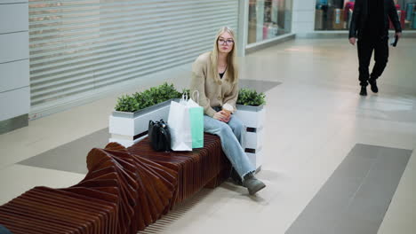 lady seated in a mall, looking pensive with a coffee cup in hand, shopping bags beside her, black handbag resting next to her, while other shoppers walk by in the background