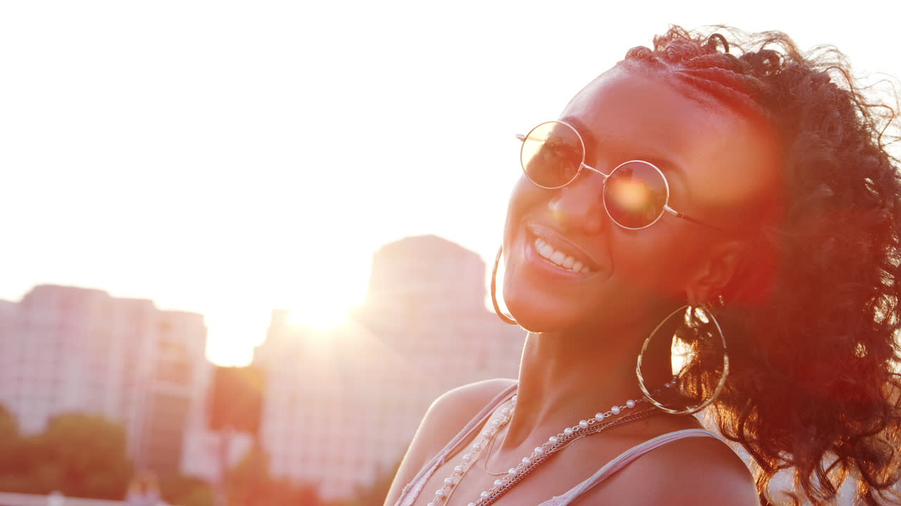 Jovem mulher negra usando óculos de sol, olhando para a câmera e rindo em uma rua de Londres na hora dourada, close-up