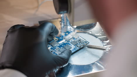 una vista trasera de las manos con guantes de un técnico trabajando en una placa de circuito azul bajo un microscopio, el técnico está usando cuidadosamente un soldador para soldar los componentes electrónicos