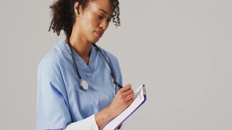 Video-of-biracial-female-doctor-writing-on-clipboard-on-blue-background