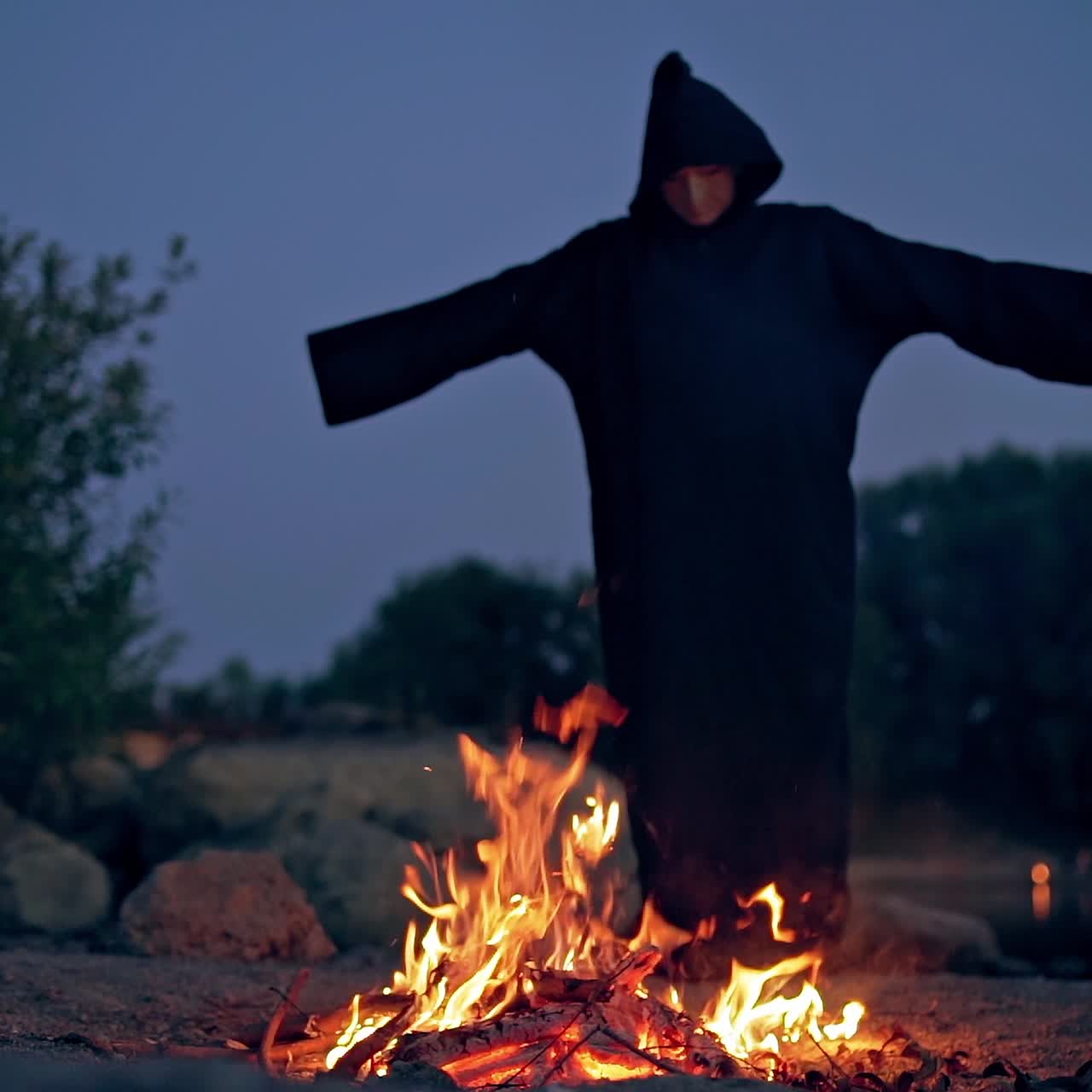 Witch in black spooky costume. Scary wizard in cloak with hood doing shamanism over the burning fire on the natural evening background. Halloween concept.