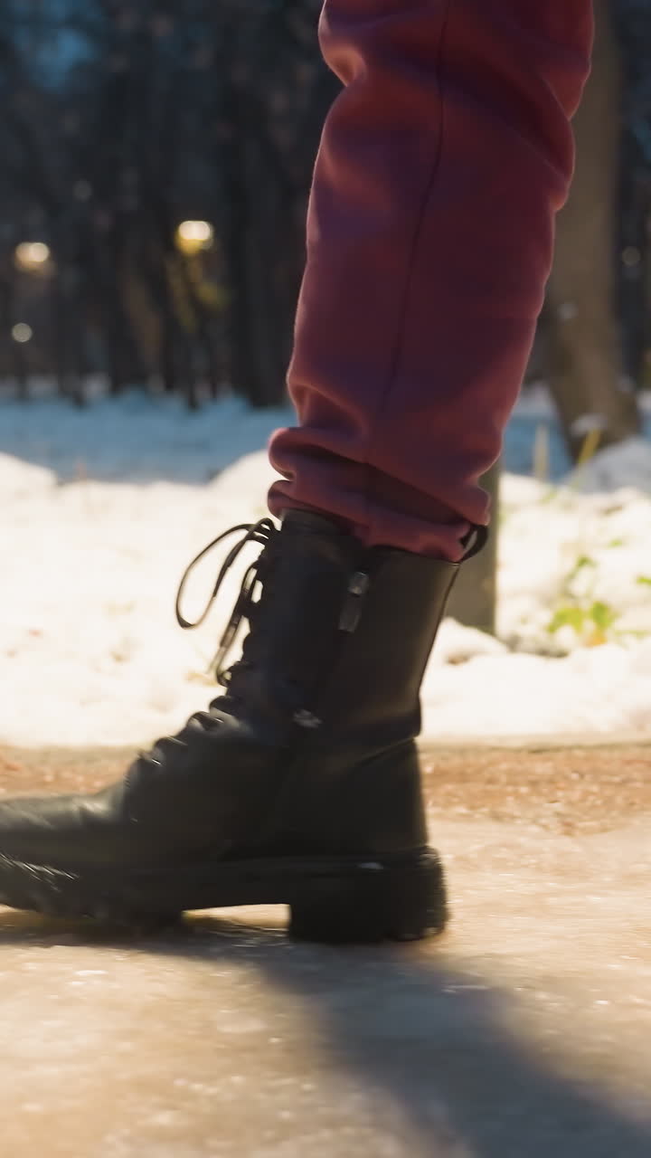 primer plano de un pie con botas pisando un camino cubierto de nieve en un parque de invierno, luces de la calle iluminadas en el fondo, mostrando nieve, clima frío y entorno invernal urbano, con suelo nevado