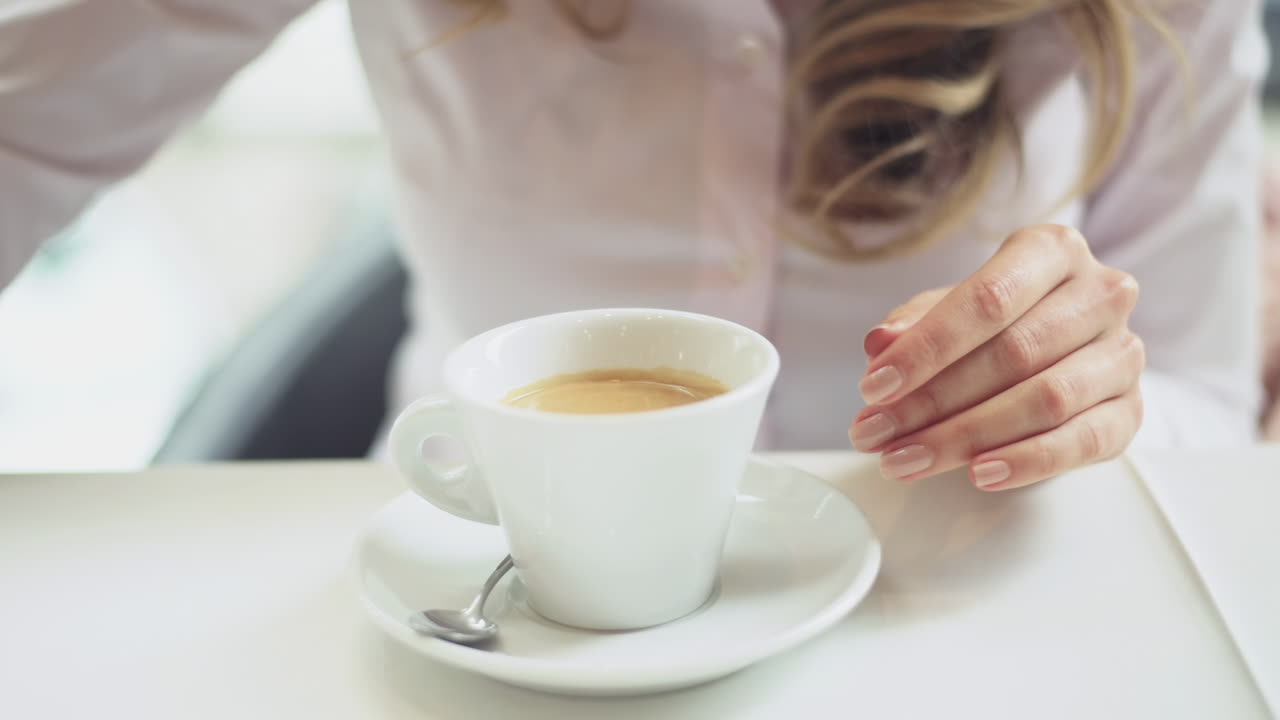 Close up of a blonde woman in a light pink shirt pouring cream and drinking coffee in front of a window