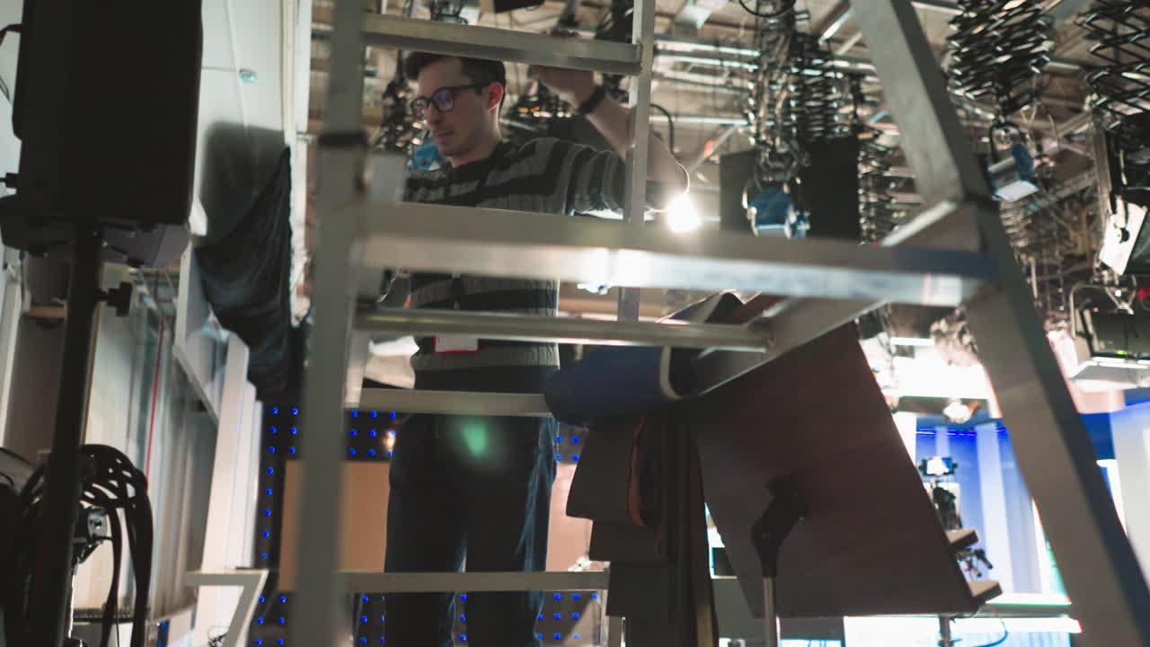 Worker climbing aluminum ladder in TV production studio to adjust lighting equipment. Industrial workspace with studio lights, cameras, and cables overhead. Behind-the-scenes broadcast setup