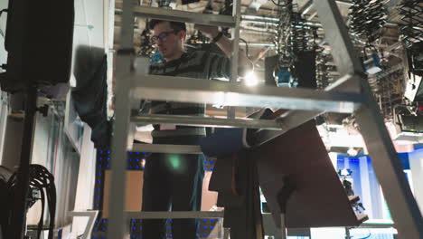 worker climbing aluminum ladder in tv production studio to adjust lighting equipment. industrial workspace with studio lights, cameras, and cables overhead. behind-the-scenes broadcast setup