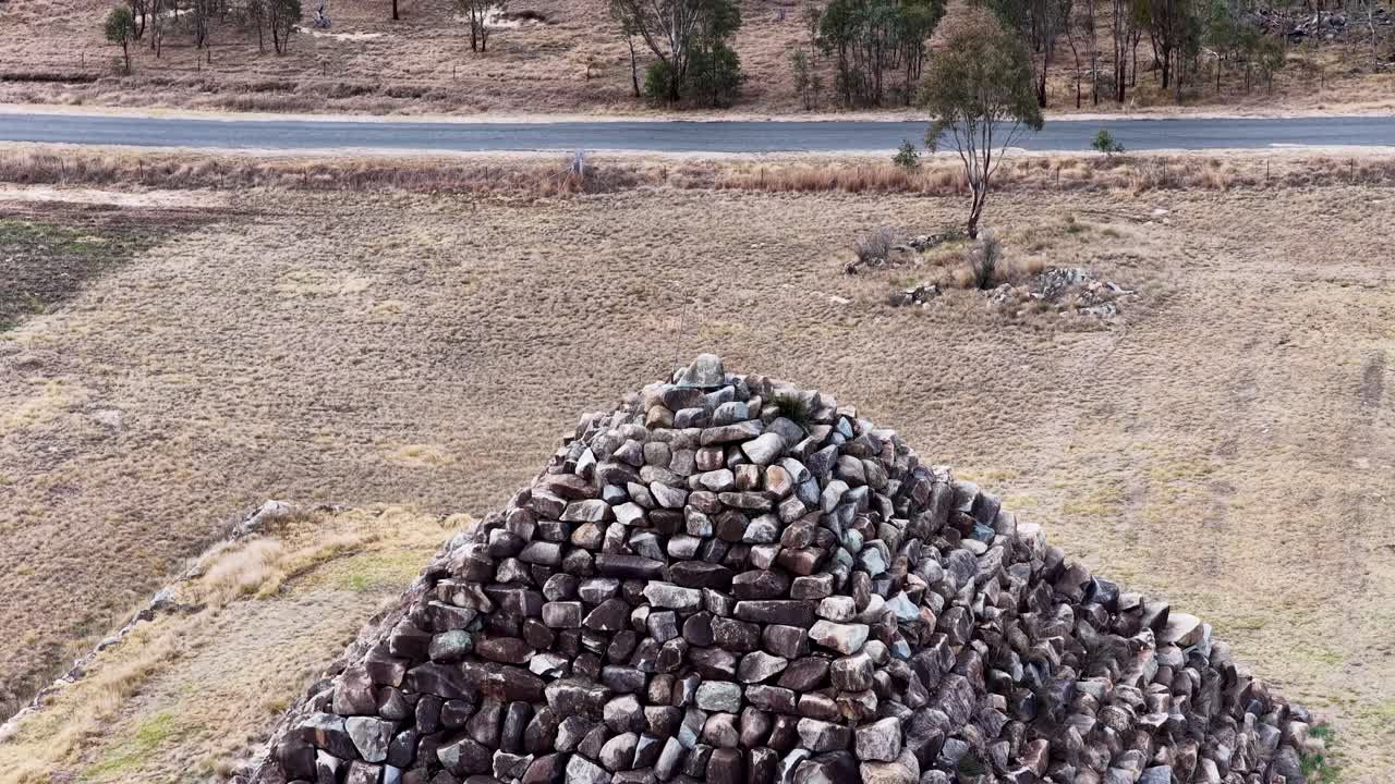 Drone camera ascends above a large granite rock pyramid in a dry, grassy field near Ballandean, Queensland, revealing surrounding bushland and rural landscape