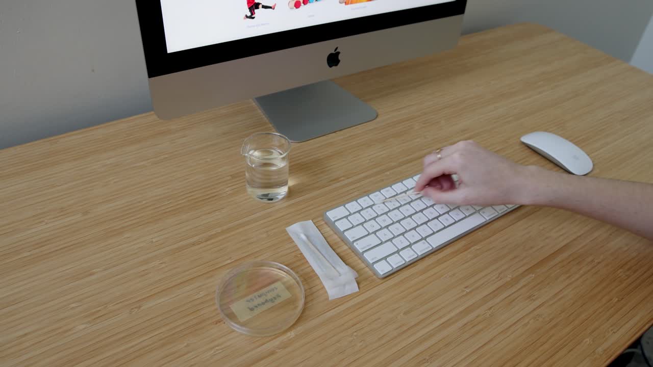 A person collects bacteria samples from a keyboard using a petri dish in a well-lit laboratory setting