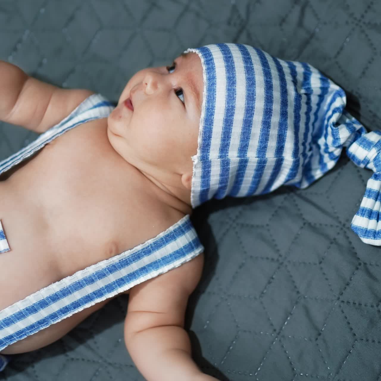 Bambino piccolo in costume da gnomo lasciato sul letto. Bambino con buffo cappello a righe che gira la testa. Primo piano. Sfondo grigio