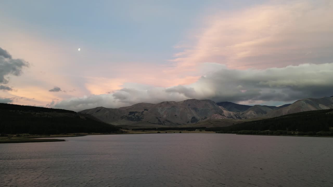 drones aéreos vuelan cerca del paisaje patagónico de la laguna de ensueño, cielo azul rosado y agua reflectante entre colinas, la luna brilla alto, lago la zeta, argentina