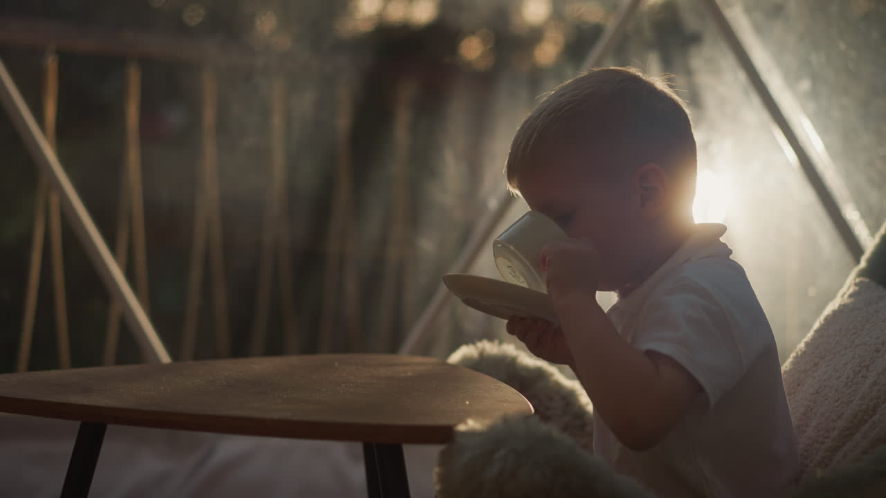아이는 글램핑에서 작은 테이블에서 차를 마시고, 어린 소년은 저녁 캠프에서 베개 위에 있는 컵에서 술 한 잔을 마시며, 테라스에서 맛있는 음료를 마신다.
